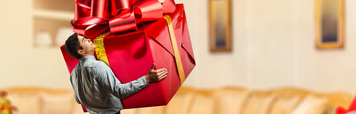 Mężczyzna niosący ogromny prezent zapakowany w czerwony papier z kokardą