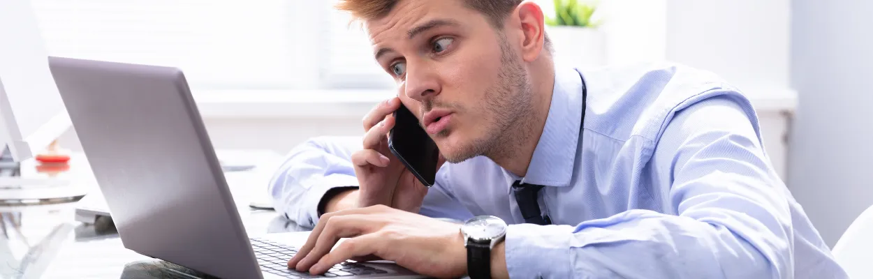 Mężczyzna rozmawiający przez telefon i siedzący przy biurku na którym znajduje się laptop